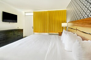 A hotel room with a large bed, a flat-screen TV mounted on the wall, a wooden headboard, a bedside table with a lamp, and a patterned wall. - Holiday Shores - ADA Standard King Room (Myrtle Beach)