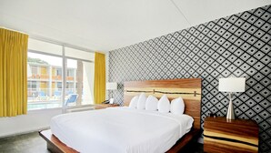 Habitación de hotel con una cama grande, cabecera de madera, mesita de noche, lámpara y vistas a una piscina y un edificio a través de una ventana.