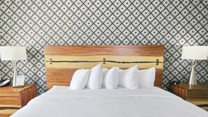 Una cama bien hecha con ropa blanca y varios almohadones, un cabecero de madera y dos mesitas de noche, una de ellas con lámpara y teléfono.