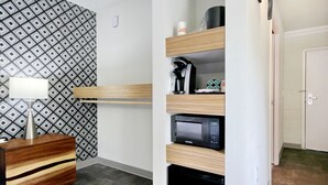Habitación de hotel con papel tapiz estampado, mesita de noche de madera con lámpara, microondas empotrado y cafetera en un estante.