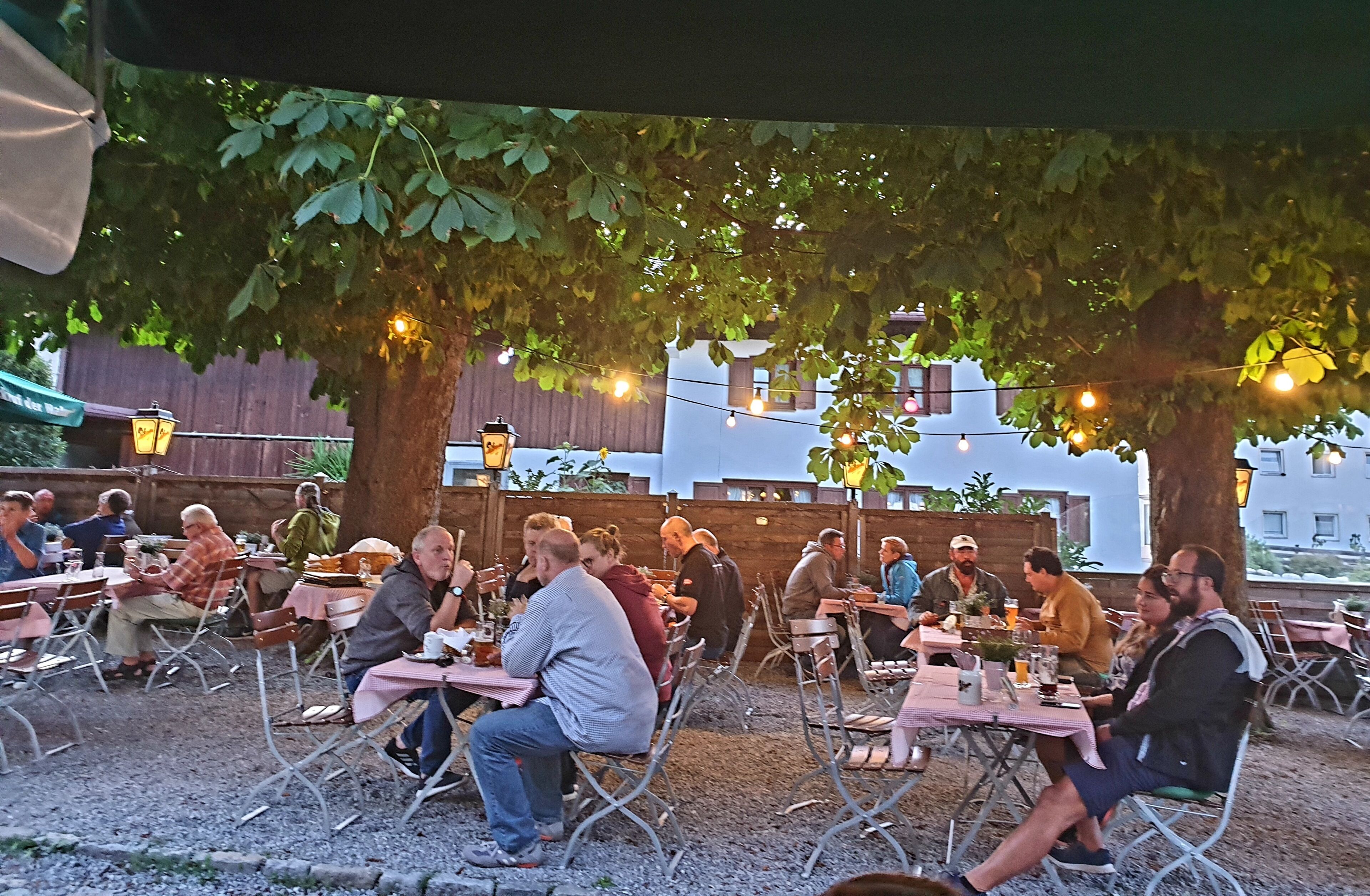 An outdoor dining area with tables and chairs under trees, string lights, and a wooden fence in the background.