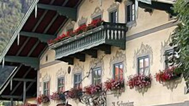 A traditional building with a sloped roof, balconies with flower boxes, and a mountain backdrop. - Ferienwohnung Typ C ca. 62 qm bis Vier Personen (Flintsbach am Inn)