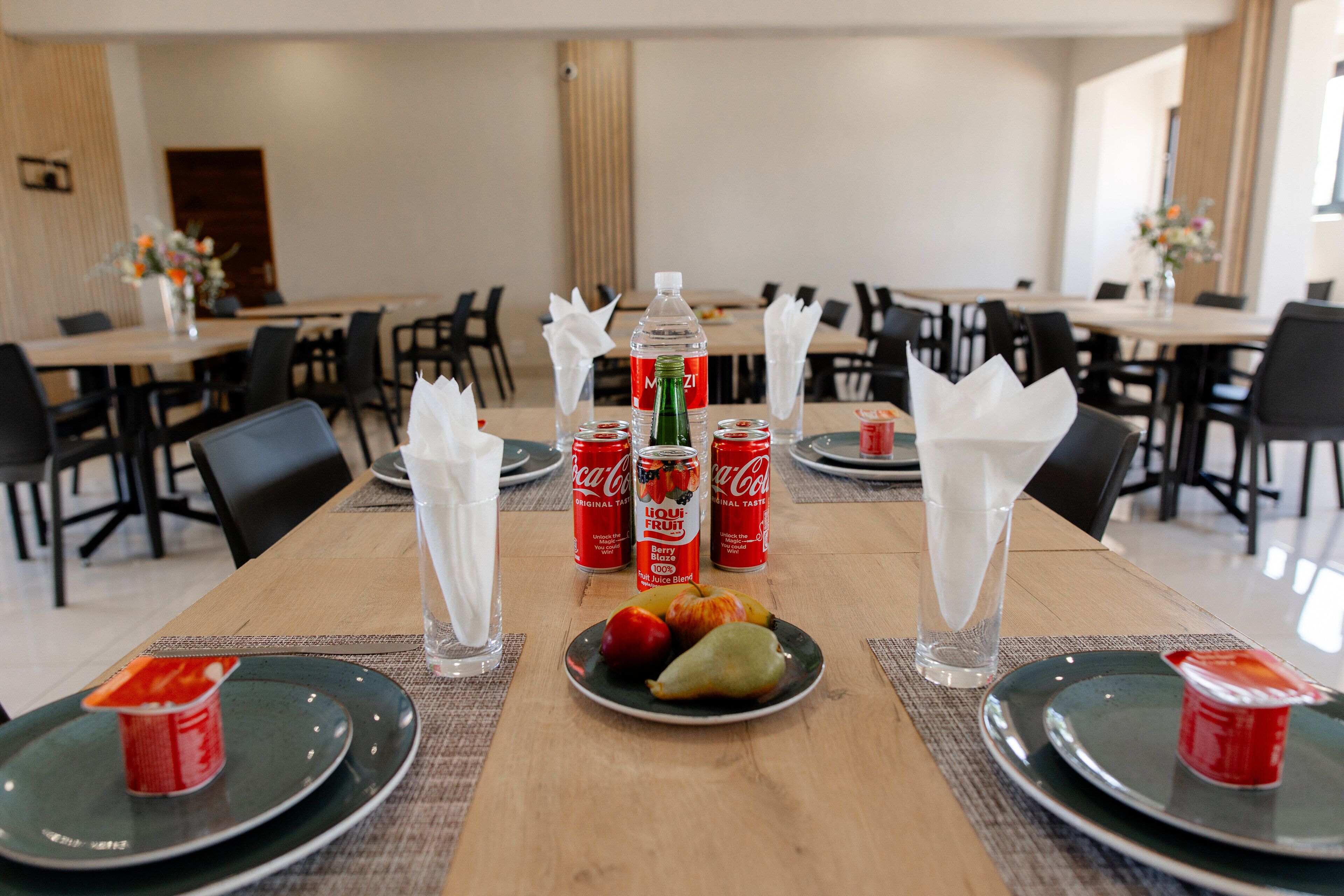 A dining area with tables set for four, each with a plate of fruit, a bottle of water, and a can of Coca-Cola.