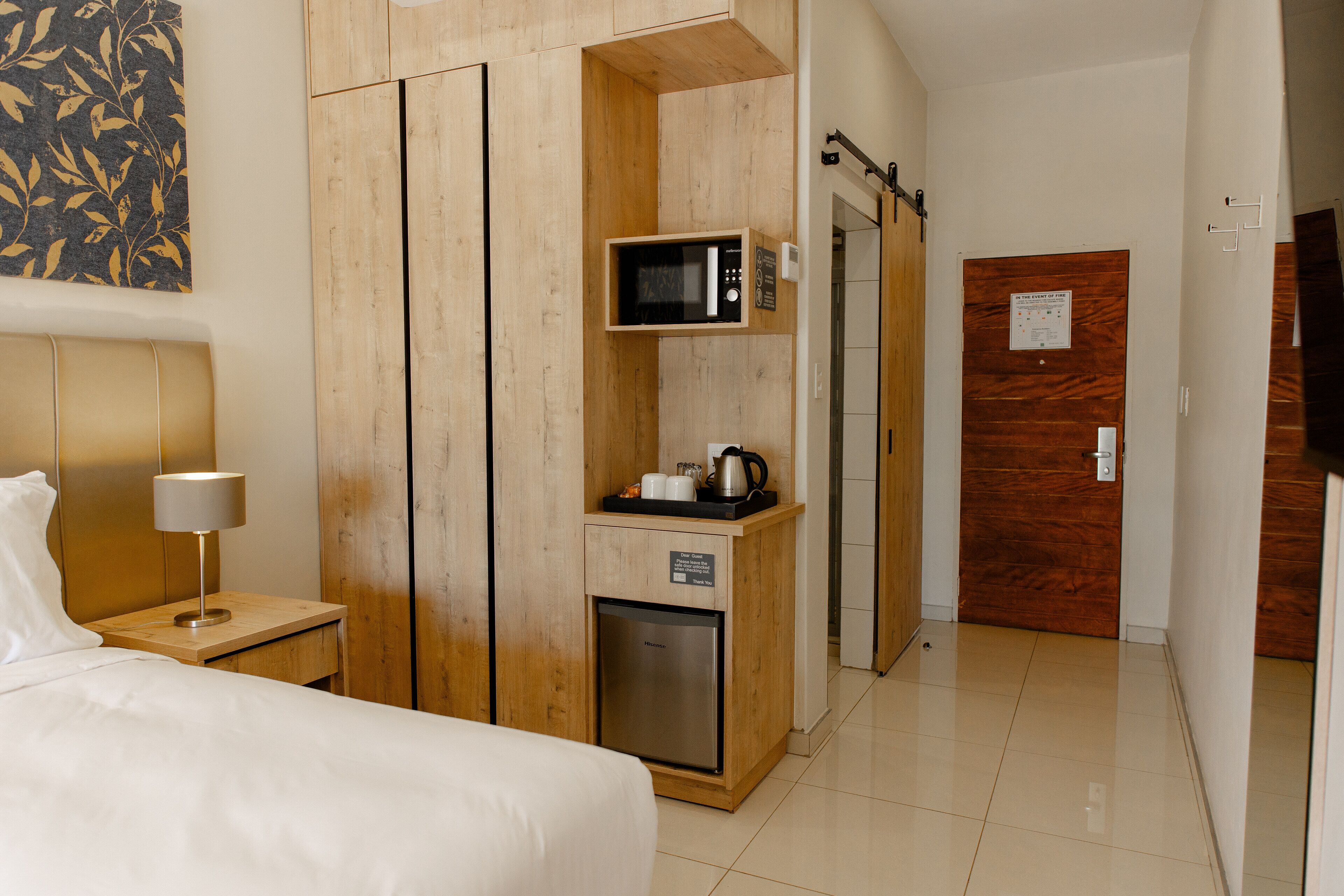 A hotel room with a bed, a nightstand with a lamp, a wooden wardrobe with a microwave and a coffee maker, and a wooden door.