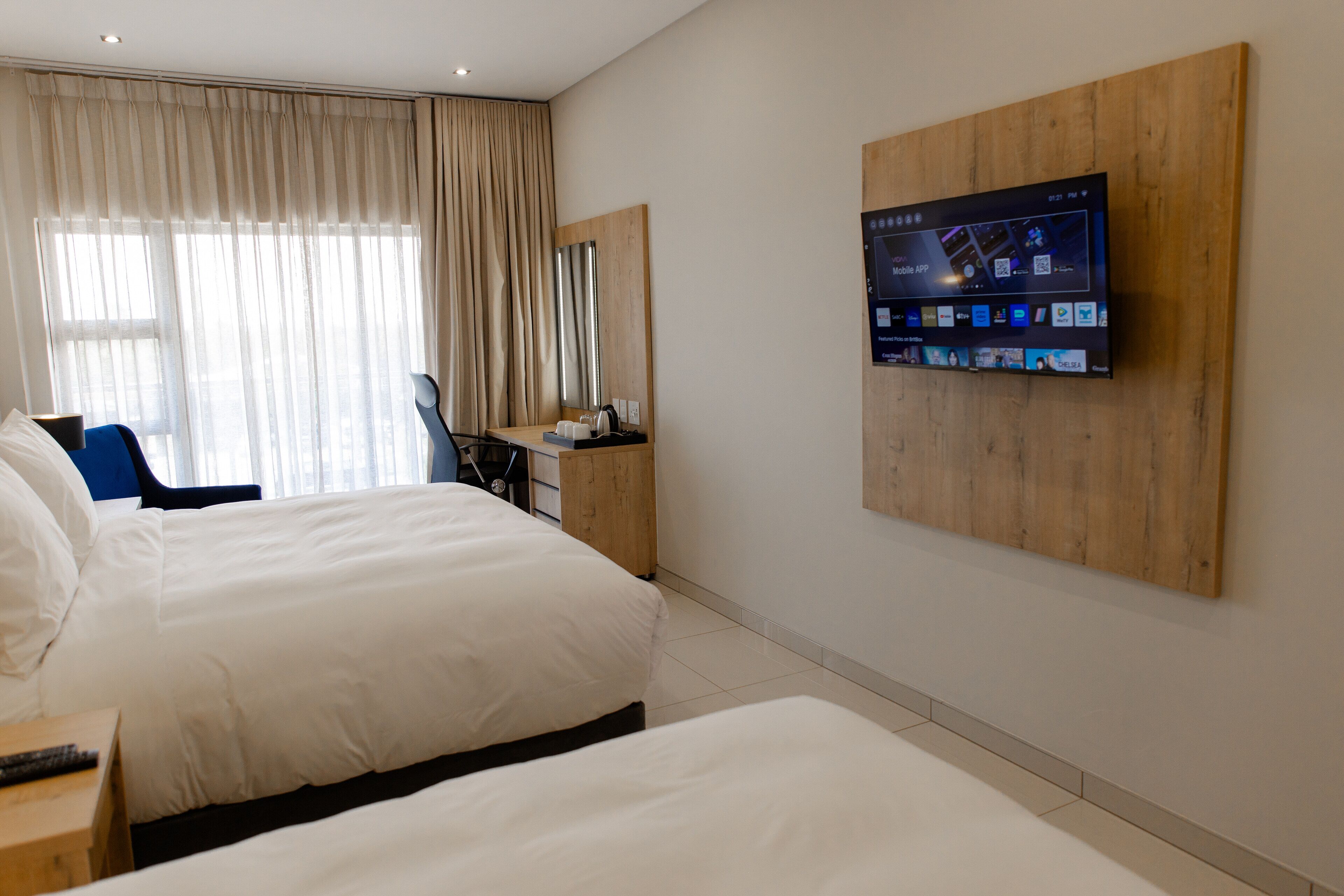 A hotel room with two beds, a desk, a chair, a TV mounted on the wall, and a large window with curtains.