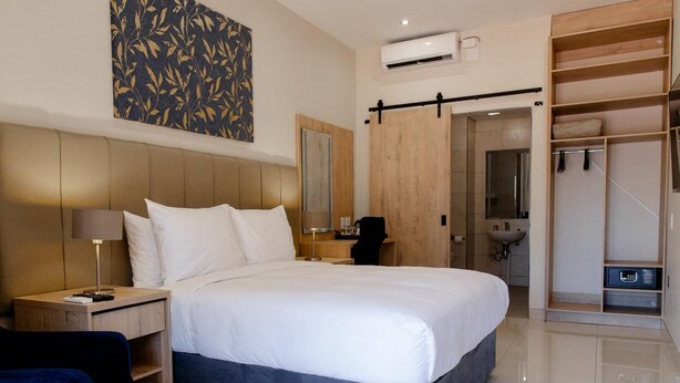 A modern hotel room with a large bed, a desk, a chair, a wardrobe, and a bathroom visible through an open door. - Acres Inn Klerksdorp (Klerksdorp)