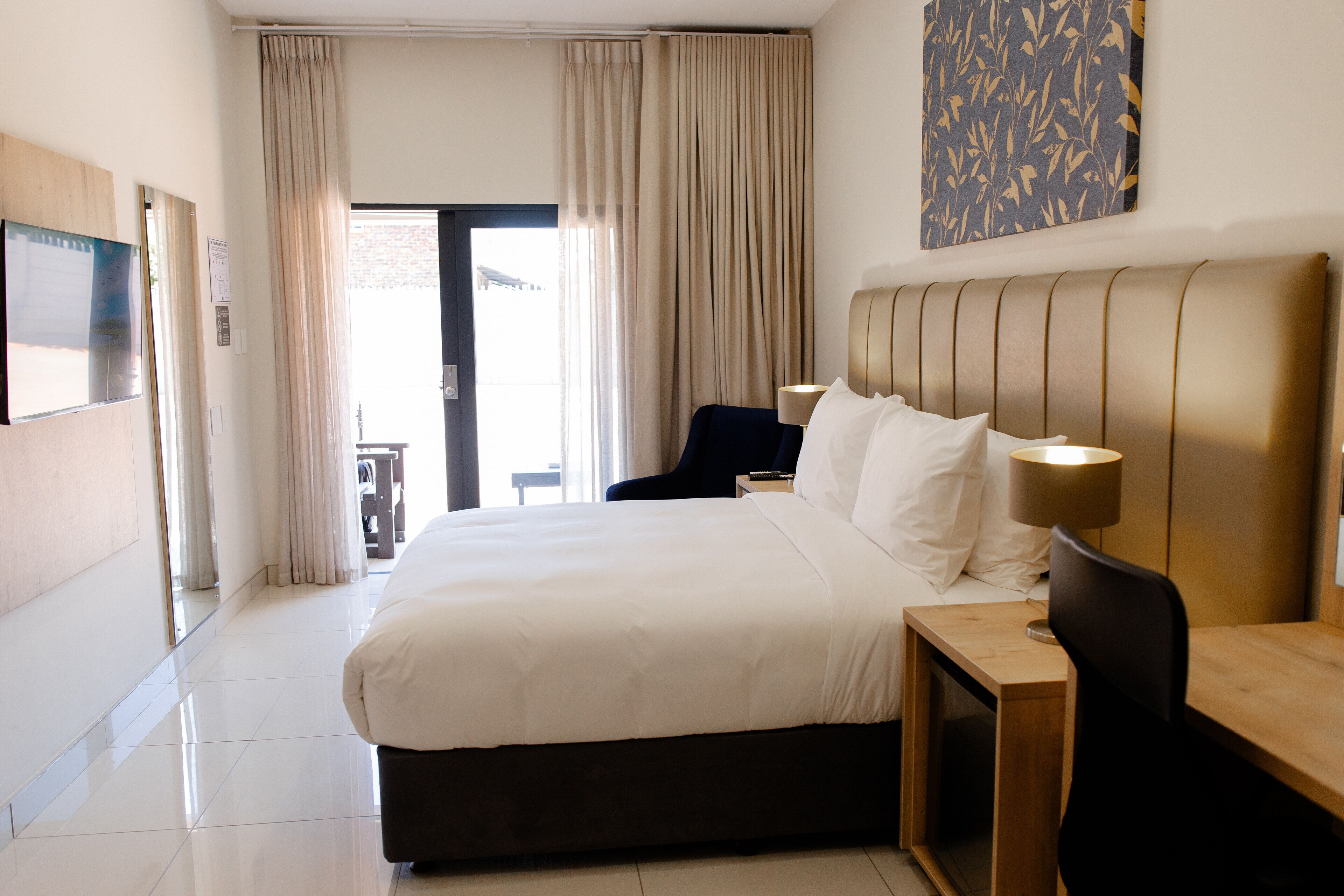 A hotel room with a large bed, a wooden headboard, two bedside lamps, and a view of the outdoors through a door.