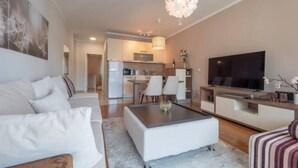Un salon moderne avec un canapé, une table basse et un téléviseur. On aperçoit une cuisine au fond.