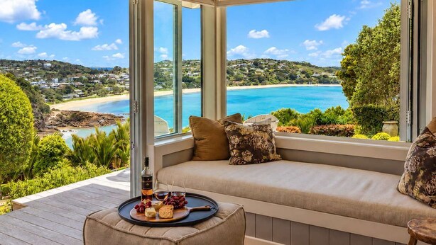 Una habitación con un gran ventanal con vistas a la playa, una zona de estar con cojines y una bandeja con comida y bebida.