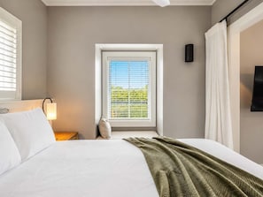 Een slaapkamer met een bed, een raam met zonwering, een bedlampje en een aan de muur bevestigde televisie.