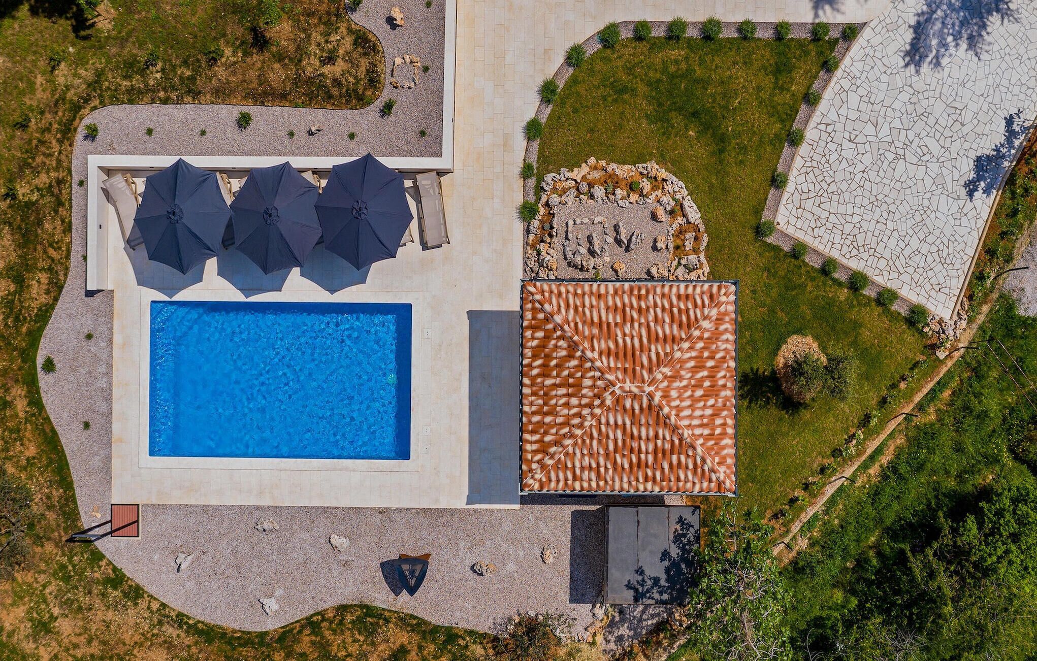 An aerial view of a pool area with a rectangular pool, a covered seating area with umbrellas, and a tiled deck.