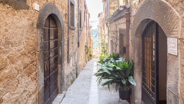 Un estrecho callejón empedrado flanqueado por edificios antiguos de piedra con portales arqueados y un letrero que indica un lugar.