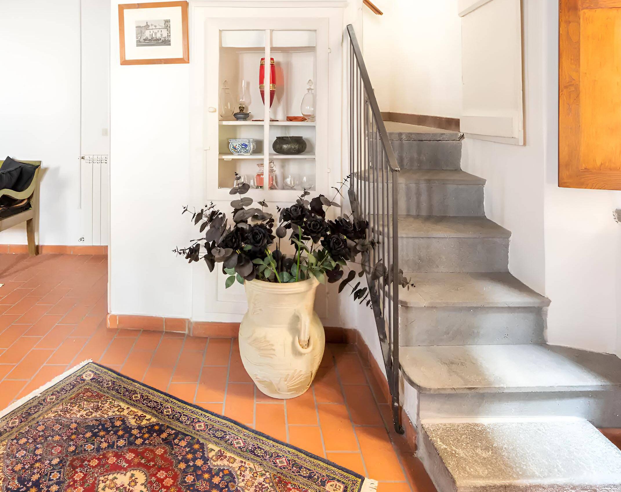 A staircase with a white railing, a white door with a framed picture, and a vase with dark flowers on the floor.