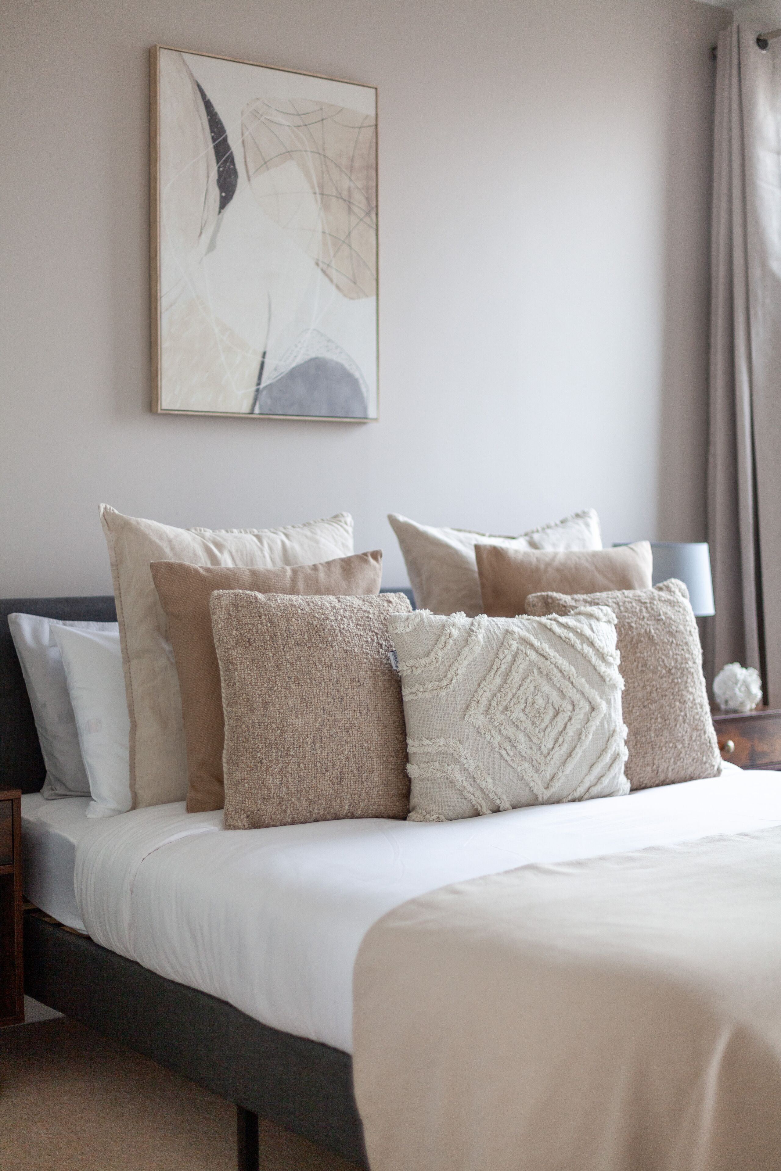 A neatly made bed with multiple pillows and a headboard, a bedside table with a lamp, and a framed abstract painting on the wall.
