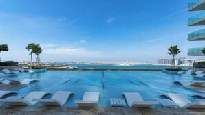 A swimming pool with lounge chairs, a clear sky, and a distant view of a coastline. - Vogue | Beach Vista near Palm Jumeirah | JBR (Dubai)