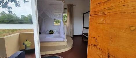 Een slaapkamer met een bed onder een hemelbed, een raam met gordijnen en uitzicht op groen.