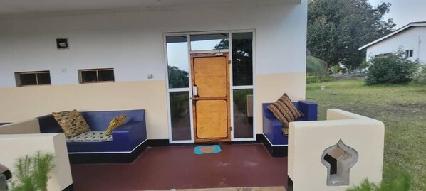A covered outdoor seating area with a sofa and cushions, a wooden door, and a small garden. - Mafia Fabu Lodge (Mafia Island)