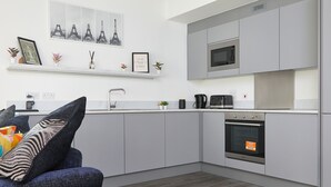 A modern kitchen with grey cabinets, a microwave, an oven, and a coffee machine. There is a blue sofa with decorative pillows and a small plant on the counter. - Immaculate 2-bed Apartment in London (London)