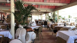 Un restaurante con terraza, mesas cubiertas con manteles blancos, sillas de mimbre y una scooter antigua.
