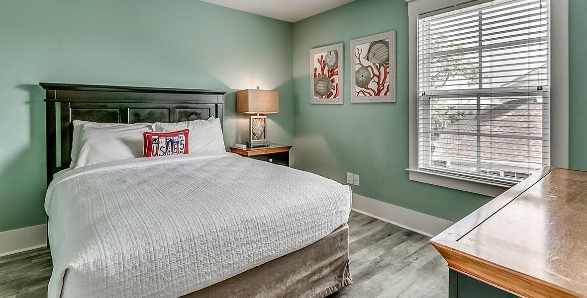A bedroom with a bed, a nightstand, a lamp, a window with blinds, and artwork on the wall.