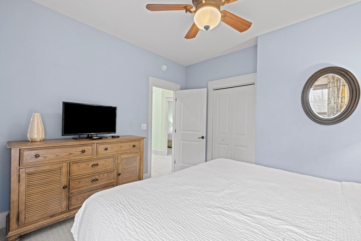 A bedroom with a bed, a wooden dresser, a television, a ceiling fan, and a door.
