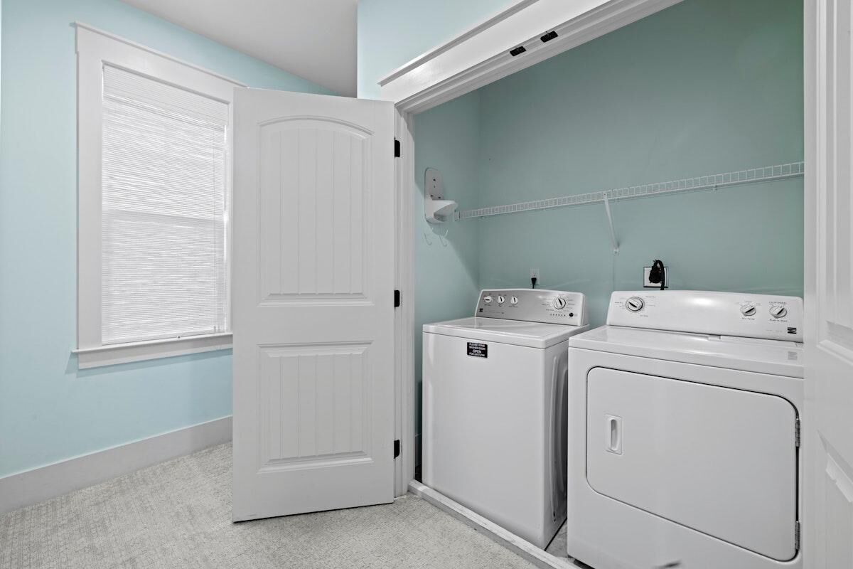 A laundry room with a white door, two stacked washers and dryers, a window with blinds, and a light blue wall.