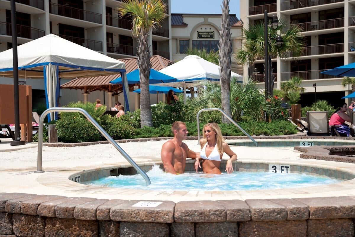 Ein Poolbereich mit Whirlpool, Palmen und im Hintergrund ein Gebäude mit der Aufschrift 'North Beach Towers'.