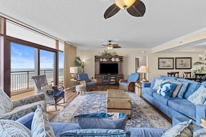 A living room with a large window overlooking the ocean, a ceiling fan, a TV, and a blue sofa.