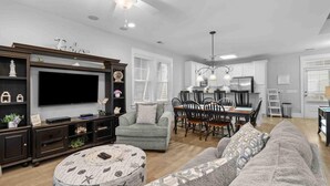 A spacious living area with a large flat-screen TV, dark wood entertainment unit, grey sofa, and a dining area with a chandelier.