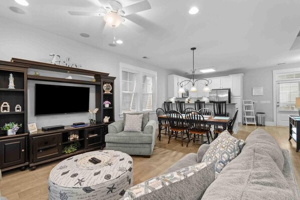 A spacious living area with a large flat-screen TV, dark wood entertainment unit, grey sofa, and a dining area with a chandelier.