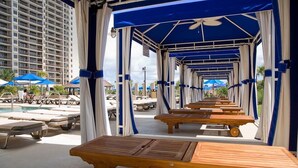 A poolside area with wooden tables and chairs, a covered seating area with blue and white curtains, and a high-rise building in the background. - Modern 1 Bedroom Luxury Flat Your Stylish Oasis (North Myrtle Beach)