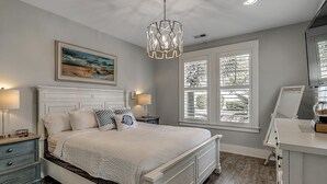A bedroom with a large bed, bedside tables, a dresser, a chandelier, and a window with blinds. - Elegant 3 Bedroom: Unmatched Comfort for 6 Guests (North Myrtle Beach)