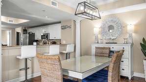 A modern kitchen with white cabinets, a stainless steel refrigerator, and a dining area with a glass table and wicker chairs. - Elegance and Comfort Await: a Perfect Retreat (North Myrtle Beach)