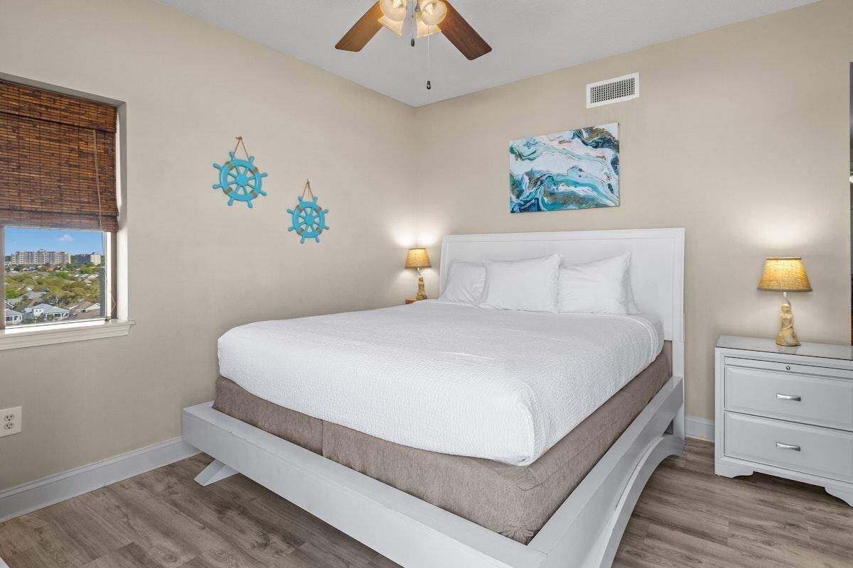 A bedroom with a bed, a ceiling fan, a nightstand, a window, and a city view.