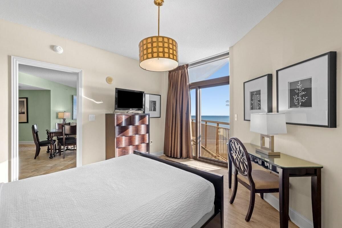 Habitación de hotel con cama, escritorio, silla, televisión y balcón con vistas.