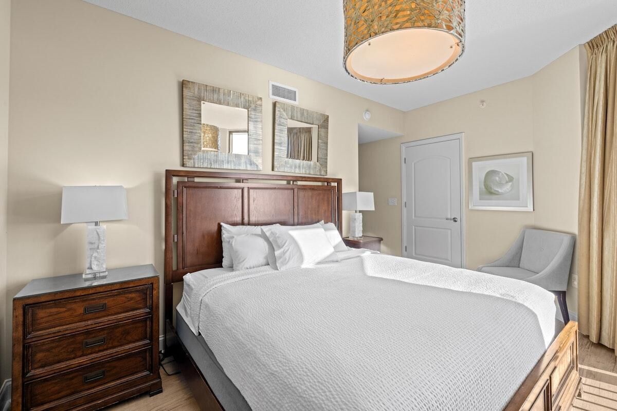 Habitación de hotel con una cama grande, cabecera de madera, dos mesitas de noche con lámparas, una silla, una puerta y un espejo en la pared.