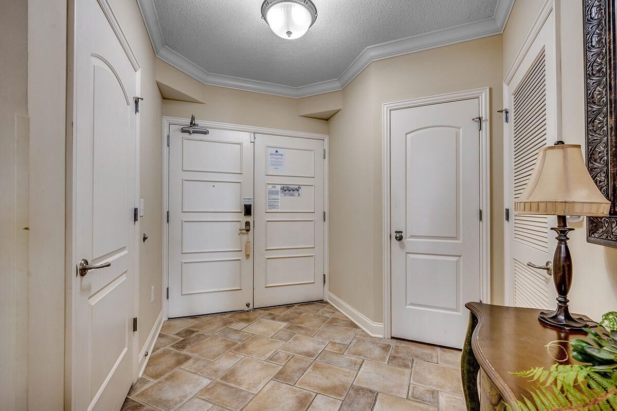 A hallway with beige walls, a tiled floor, and white double doors. There is a lamp on a wooden table to the right.