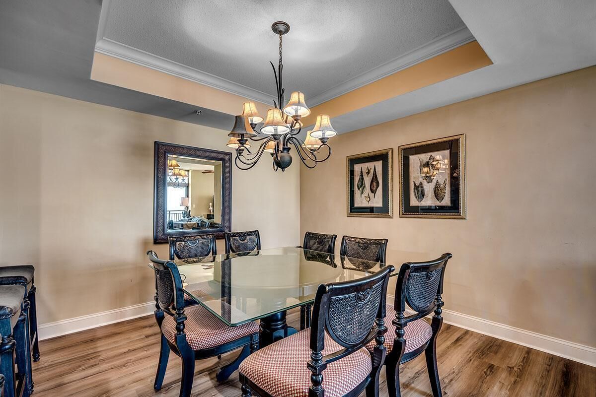 A dining room with a glass table surrounded by six chairs, a chandelier, and two framed artworks on the walls.