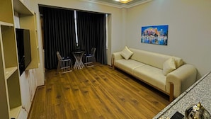 A modern living room with a sofa, a dining table with chairs, a flat-screen TV mounted on the wall, and a painting of a cityscape.
