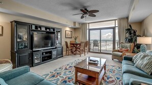 A spacious living room with a large flat-screen TV, a ceiling fan, and a balcony with a view. - Indigo Tower: Luxurious Oceanfront 4 Bdrm Paradise (North Myrtle Beach)