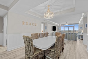 Un coin repas avec une table blanche et des chaises en osier, un lustre et une vue sur l’océan.