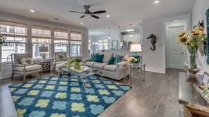 A spacious living room with a large blue rug, a ceiling fan, and a dining area with a wooden table and chairs. - Coastal Dream: 4 Bdrm Vacation Home with Pool (North Myrtle Beach)