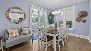 Un coin repas avec une table en bois et des chaises blanches, un canapé gris avec des coussins à carreaux et un grand miroir au mur.