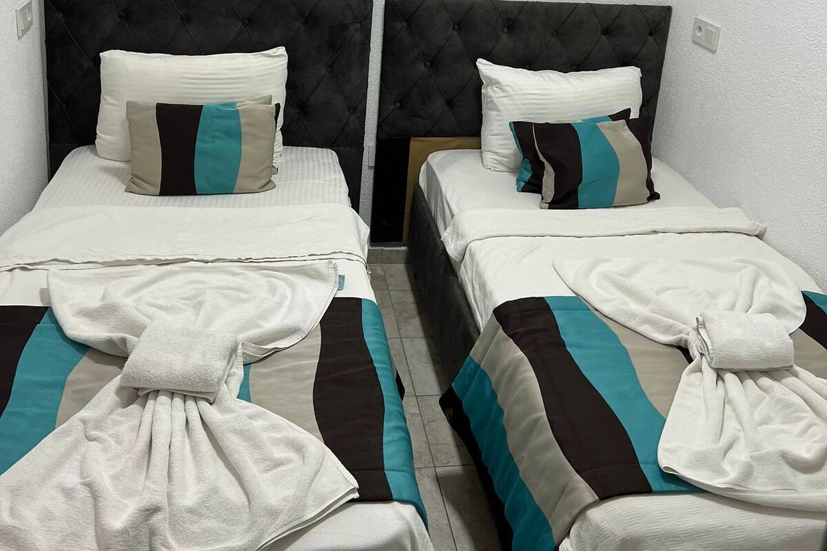 Une chambre d’hôtel avec deux lits, chacun doté d’une tête de lit capitonnée, de draps blancs et de couvertures rayées dans les tons turquoise et marron. Un seul luminaire est suspendu au plafond.