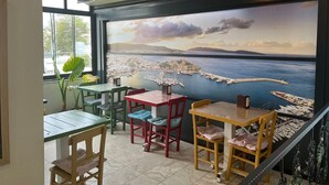 A dining area with tables and chairs, a large mural of a coastal scene, and a view of the sea through large windows. - Tiana Butik Otel (Bodrum)