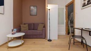 Una habitación pequeña y acogedora con un sofá morado, una mesa redonda blanca, una lámpara de pie y una puerta de madera.