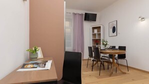 Una habitación moderna con un escritorio de madera, sillas negras, una mesa redonda, un televisor montado en la pared y una estantería.