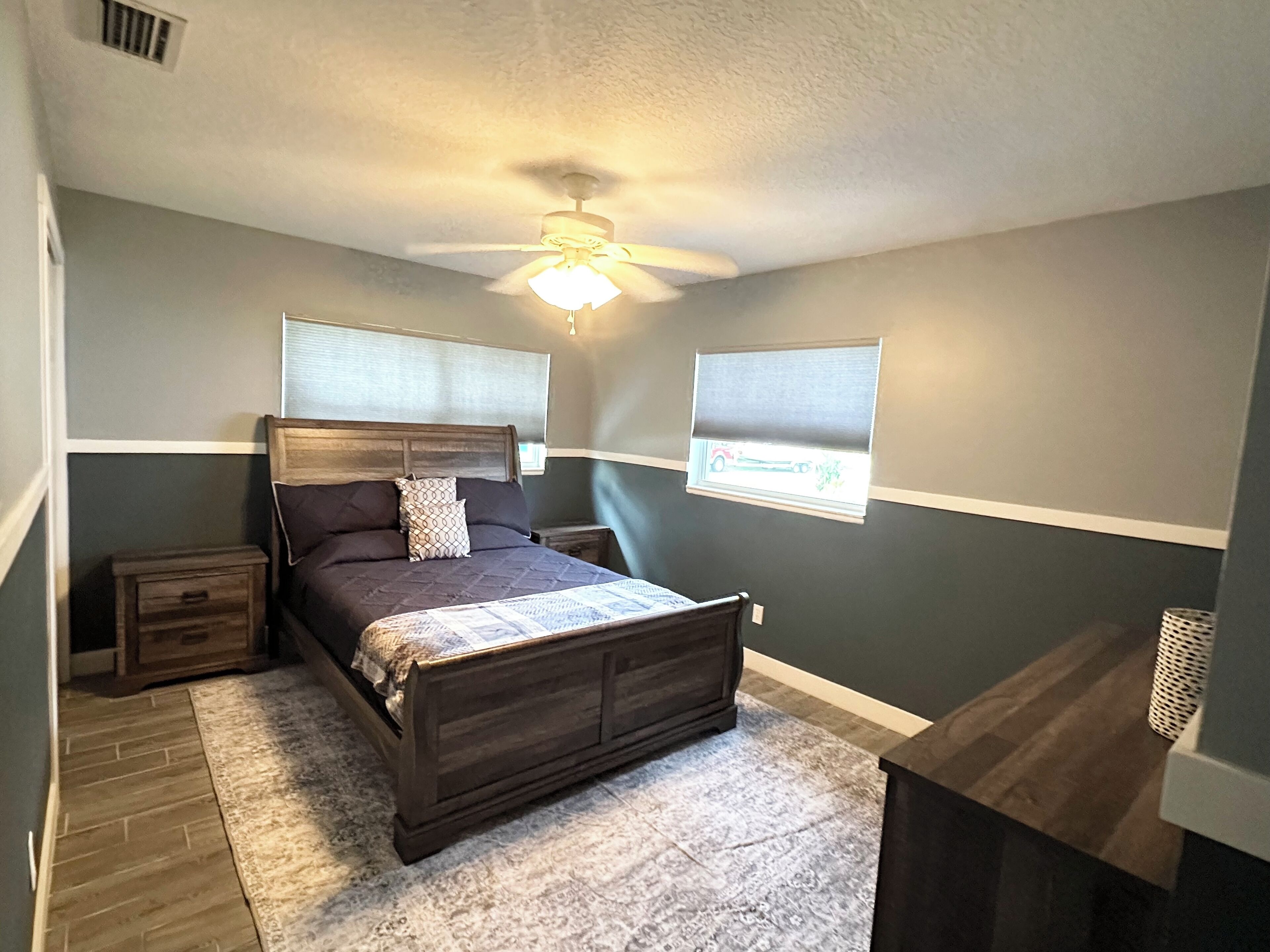 A bedroom with a bed, a nightstand, a ceiling fan, and two windows with blinds.