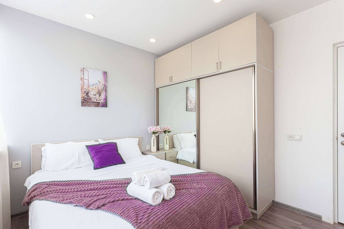 A modern hotel room with a large bed, a wardrobe, a mirror, and a painting on the wall.