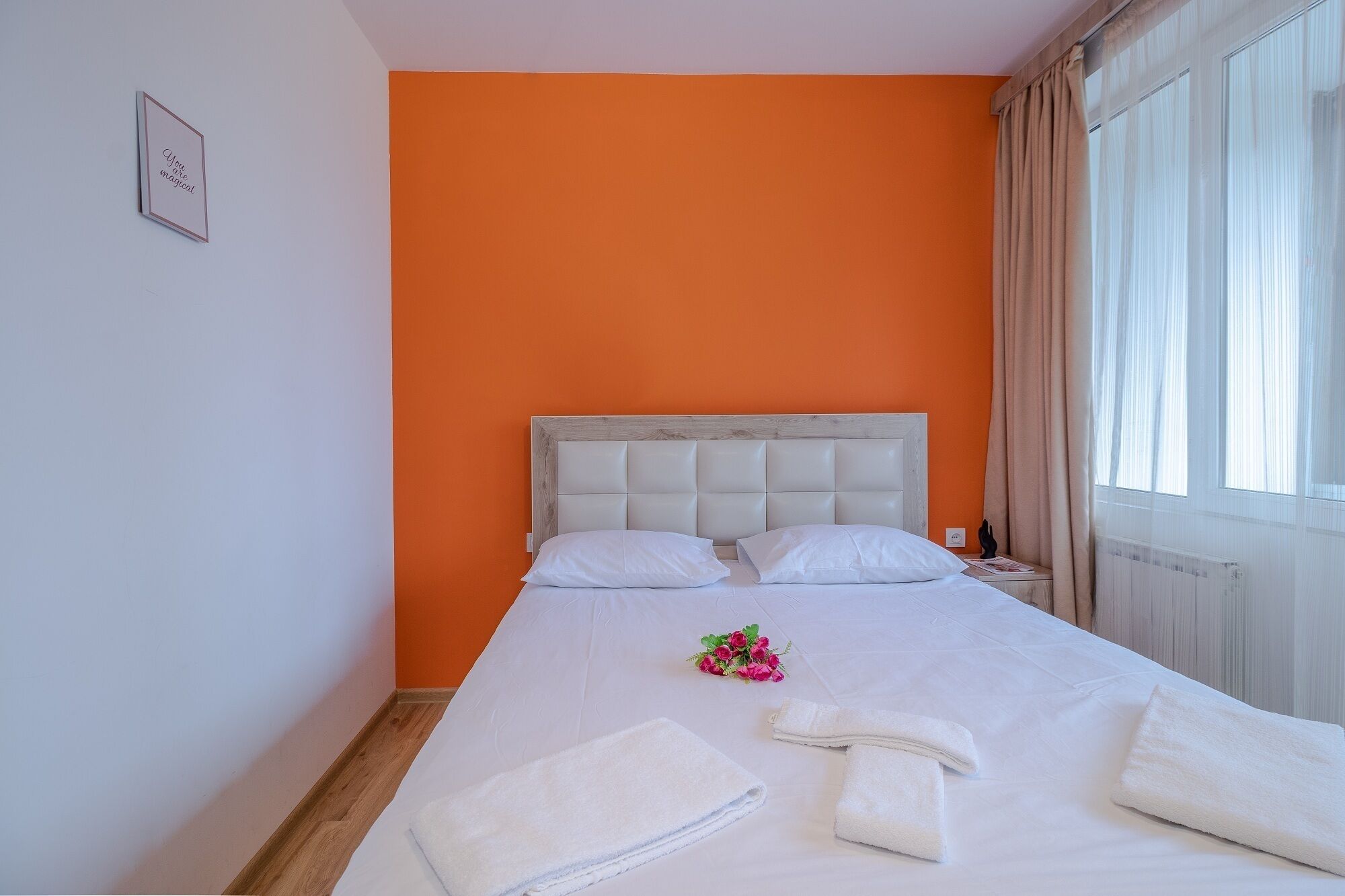 Een hotelkamer met een oranje accentmuur, een wit bed met twee kussens, een bloemenboeket en netjes opgevouwen handdoeken op het bed.
